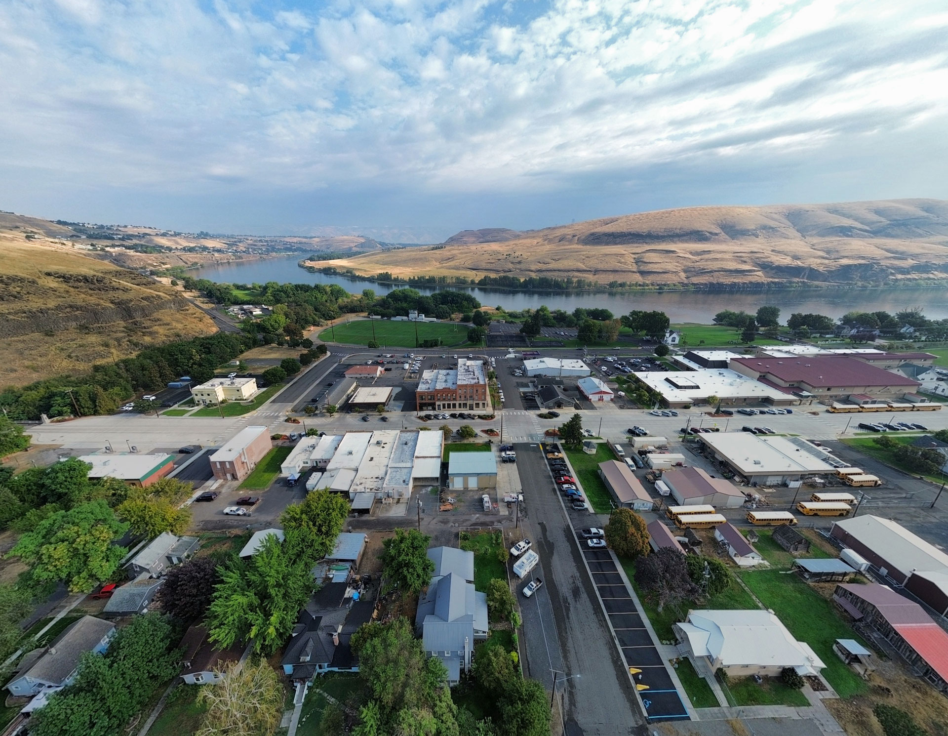 Aerial view of Asotin, WA, with houses, buildings, and streets nestled among green trees and hills. A wide river flows in the background, bordered by rolling hills under a partly cloudy sky.