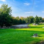 A lush green park with benches, picnic tables, a parking lot, and tall trees under a blue sky with scattered clouds. A single car is parked on the lot, and hills appear in the background. Asotin, WA