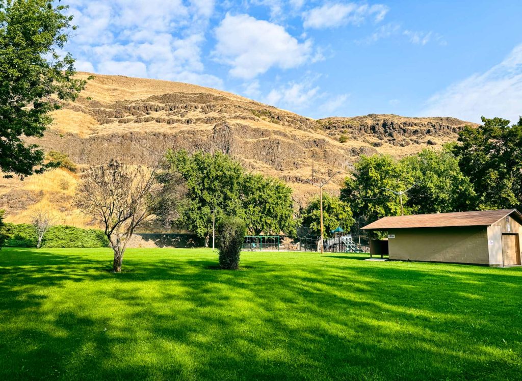 A small tan building sits on a lush green lawn, bordered by leafy trees. In the background, steep, rocky hills with dry yellow grass rise under a partly cloudy blue sky.