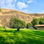 A small tan building sits on a lush green lawn, bordered by leafy trees. In the background, steep, rocky hills with dry yellow grass rise under a partly cloudy blue sky.