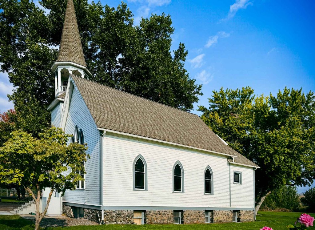 A white wooden church with arched windows and a tall steeple stands surrounded by green trees under a bright blue sky in Asotin, WA. The building sits on a stone foundation, with pink flowers visible in the foreground.
