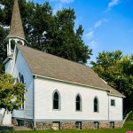 A white wooden church with arched windows and a tall steeple stands surrounded by green trees under a bright blue sky in Asotin, WA. The building sits on a stone foundation, with pink flowers visible in the foreground.