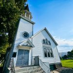 A white wooden church with arched windows and a steeple stands under a clear blue sky in Asotin, WA, surrounded by green grass and trees. Steps lead up to double doors at the entrance.