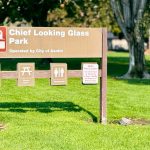 A sign for Chief Looking Glass Park in Asotin, WA stands on green grass, displaying icons for a watercraft launch, picnic area, and restrooms. Trees and a fence are visible in the background.
