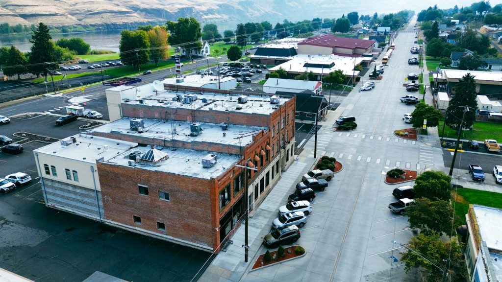 Aerial view of Asotin, WA, featuring brick buildings, parked cars, tree-lined streets, and a river with hills in the background. Streets appear clean and orderly on a clear day.
