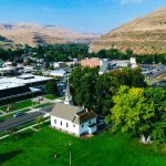 Aerial view of Asotin, WA, with a mix of buildings, roads, trees, and a white church on green grass; hills covered in dry grass rise in the background under a blue sky.