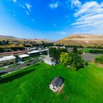 Aerial view of Asotin, WA, a small town surrounded by grassy fields and hills, with a few buildings, trees, and a bright blue sky with scattered clouds overhead.