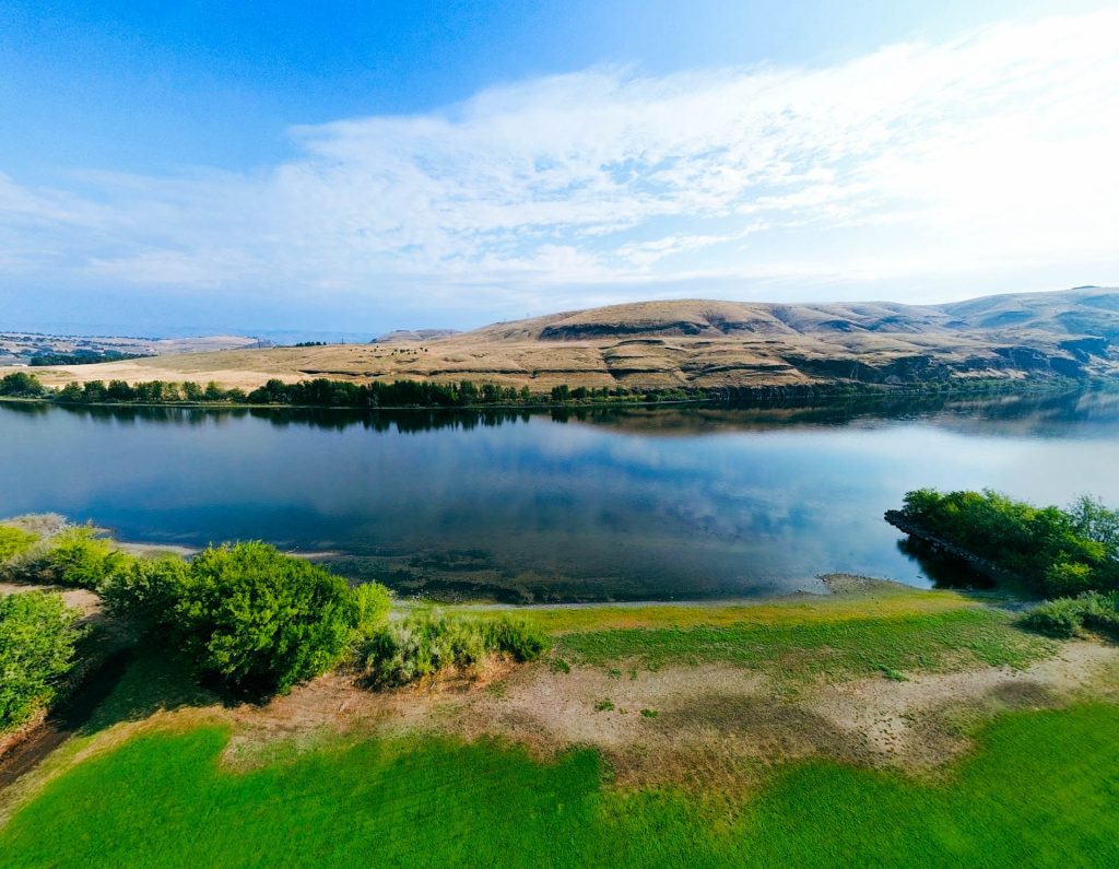 A scenic view of a calm river reflecting the blue sky and clouds in Asotin, WA, bordered by green grass and bushes, with dry, rolling hills in the background under a partly cloudy sky.