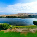 A scenic view of a calm river reflecting the blue sky and clouds in Asotin, WA, bordered by green grass and bushes, with dry, rolling hills in the background under a partly cloudy sky.