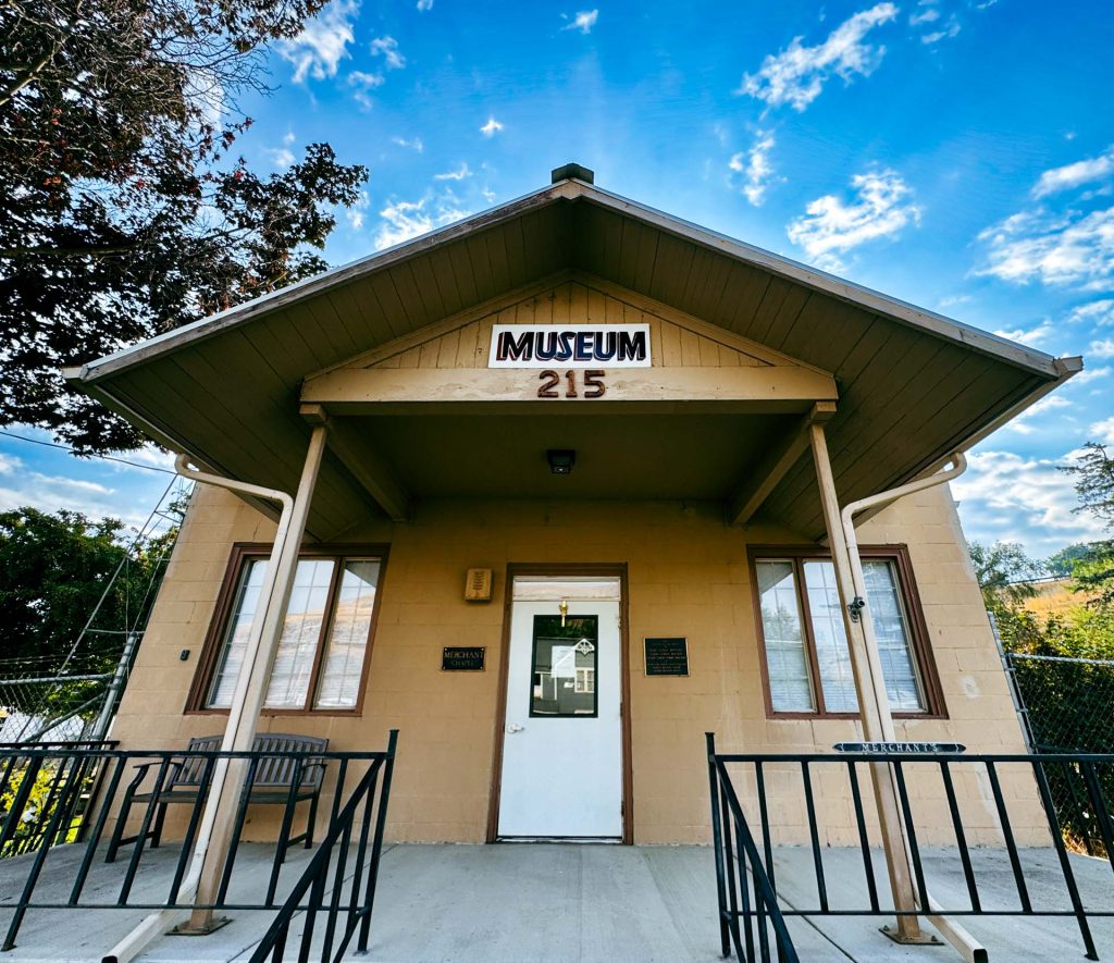 A small beige building with a front porch, labeled Museum 215 above the entrance, sits under a blue sky with scattered clouds. Black railings and benches are on either side of the white door. Asotin, WA