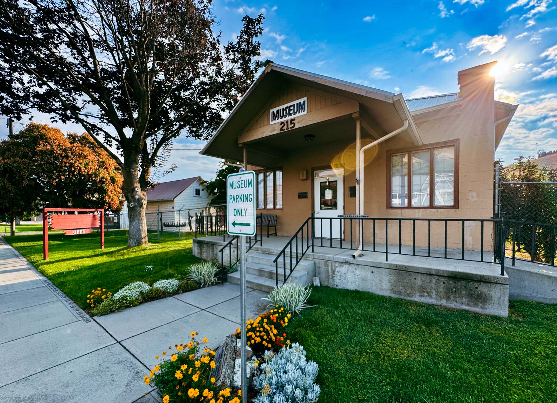 A small tan building with a porch and a sign reading Museum 215. A Museum Parking Only sign is in front, surrounded by flowers and green grass, with trees and a red sign visible nearby under a blue sky. Asotin, WA